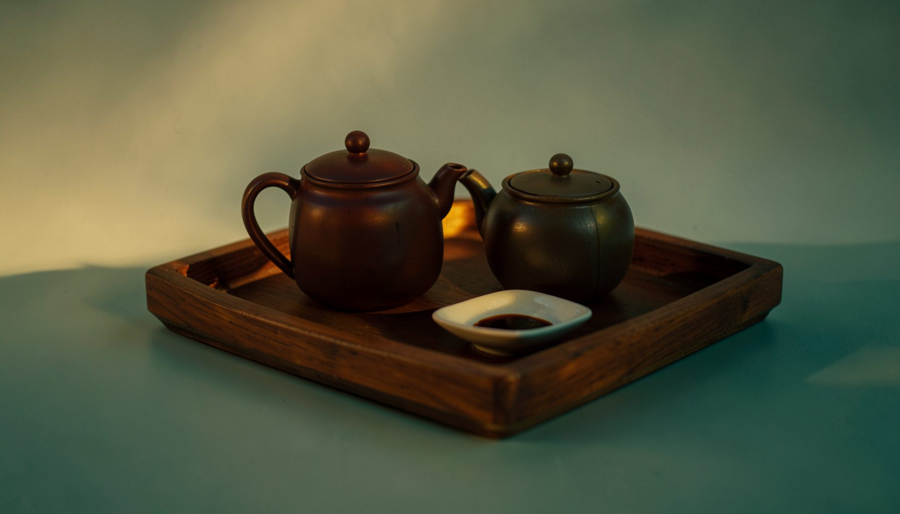 Wooden tray with a teapot and small dish, bathed in amber late-afternoon light against a muted sage background.